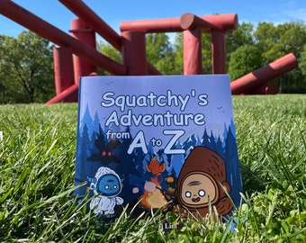 May include: A blue book with the title "Squatchy's Adventure from A to Z" on a green grassy field with a red metal sculpture in the background.