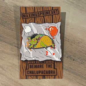May include: A green monster with red eyes is inside a yellow taco. The taco is on a white paper with red sauce and a white napkin. The text on the card reads "Beware the ChalupaCabra" and "MaximusPrimeArt".