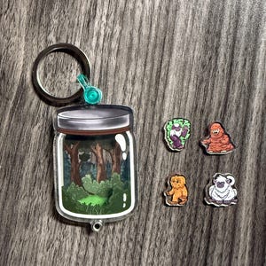 Pode incluir: Um chaveiro de acrílico transparente com uma cena de floresta dentro de um frasco, com um porta-chaves prateado e um detalhe turquesa. Quatro pequenos pingentes de criaturas coloridas também estão presentes.