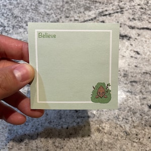Peut inclure: Une carte de vœux verte avec une bordure blanche et le mot "Believe" imprimé en vert. Un personnage de dessin animé se cache derrière un buisson en bas à droite.