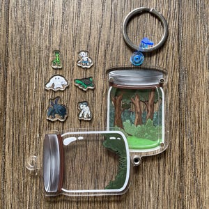 May include: A clear acrylic keychain featuring a jar with a forest scene inside, a separate jar, and several small character charms. The keychain has a silver ring and a blue accent. The charms include an alien, a turtle, and other creatures.