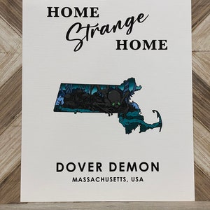 May include: A white print with black text that reads "Home Strange Home" and "Dover Demon Massachusetts, USA". The print features a blue and green illustration of the state of Massachusetts with a black silhouette of a creature with multiple eyes.
