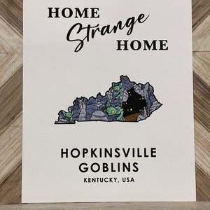 May include: A white print with black text that reads "Home Strange Home" and "Hopkinsville Goblins Kentucky, USA". The print features a cartoon illustration of a cave with green goblins inside, shaped like the state of Kentucky.