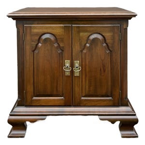May include: A dark brown wooden cabinet with two doors and brass handles. The cabinet has a rectangular shape with a flat top and decorative carved details on the doors and base. The doors have a curved design at the top.