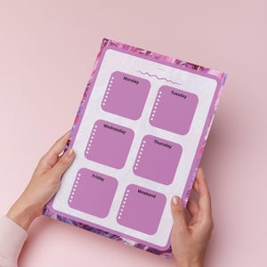 Può includere: Un pianificatore settimanale con interno bianco e bordo viola, tenuto in mano su sfondo rosa. Il pianificatore ha sei caselle viola con le etichette Lunedì, Martedì, Mercoledì, Giovedì, Venerdì e Weekend. Ogni casella ha una linea tratteggiata.