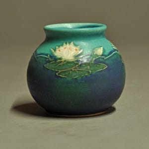 May include: A small, round ceramic vase featuring a gradient of blue and teal colors. The surface is decorated with a raised, white water lily flower and green lily pads that wrap around the body of the vessel, creating a textured relief.