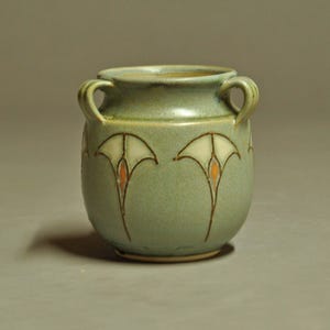 May include: A sage green ceramic vessel featuring two small side handles and a wide circular opening. The exterior is decorated with a repeating pattern of stylized white floral motifs, each accented with a small orange center and dark brown outlines.