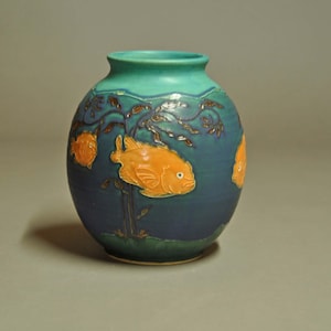 May include: A ceramic vase with a teal rim and a dark blue body. The vase features orange fish and aquatic plant designs. The fish are raised and have detailed fins. The vase has a rounded shape and a smooth finish.