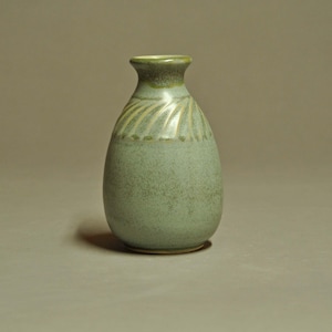 May include: A small, bulbous ceramic vase with a narrow neck and a flared rim. The body features a speckled sage green glaze, while the upper shoulder is decorated with a series of carved, radiating lines that transition into a darker green tone.