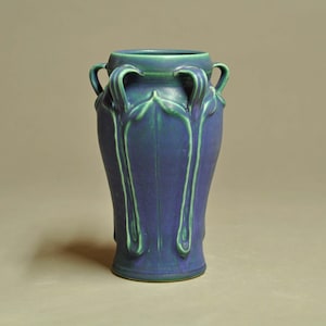 May include: A ceramic vase featuring a deep blue body with light green accents along the rim and handles. The vessel has a cylindrical shape with four symmetrical handles extending from the neck, decorated with raised vertical leaf-like motifs.