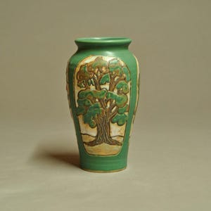 May include: A green ceramic vase featuring a central embossed panel with a detailed brown and green tree design. The vase has a smooth matte finish, a rounded body, and a flared rim, showcasing a relief illustration of a tree on a light background.