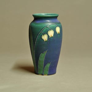 May include: A ceramic vase with a tapered neck and a smooth finish, featuring a deep blue and green gradient background. The surface is decorated with a raised relief design of three white bell-shaped flowers on green stems and leaves.