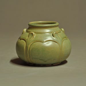 May include: A small, round ceramic vase with a sage green glaze and a smooth, matte finish. The vessel features a wide, circular opening at the top and a series of raised, petal-like decorative ridges that extend vertically around the body.