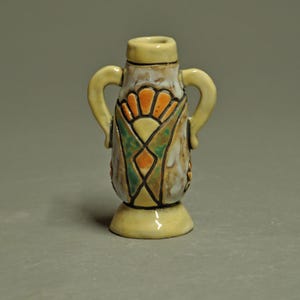 May include: A small, ceramic vase with a cream-colored base, neck, and handles. The body features a geometric design with orange, green, and yellow accents. The vase is approximately 4 inches tall, with a decorative, handcrafted appearance.