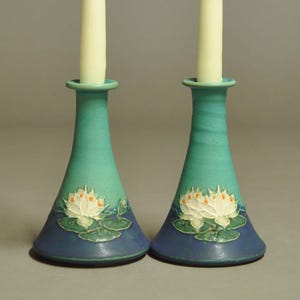 May include: A pair of teal ceramic candle holders with a flared base and a slender neck. Each holder features a white and orange water lily design with green leaves. The candle holders are designed to hold a single white candle.