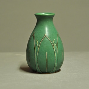 May include: A green ceramic vase with a bulbous body and a narrow neck. The vase features a stylized floral design in a lighter green and tan color. The vase is approximately 6 inches tall.