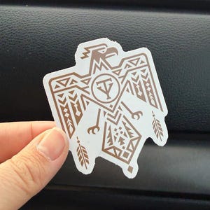 May include: A white sticker with a brown Thunderbird design. The Thunderbird has geometric patterns and a circular emblem with the letter "Y" in the center. The sticker is held by a hand.