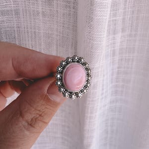 May include: A silver ring with a large, oval, light pink stone. The stone is surrounded by a detailed, beaded silver setting. The ring is held up against a white background.