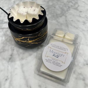 Puede incluir: Un recipiente de plástico transparente con dos fundidos de cera blanca en su interior. La etiqueta dice "SCOTLIN SCENTS WWWSCOTLINSCENTS.COM Moonlight Path NET WT: 2.5 OZ Top Note - Violet, Lavender Middle Note - Vanilla, Jasmine, Amber Bottom Note - Musk, Sandalwood"