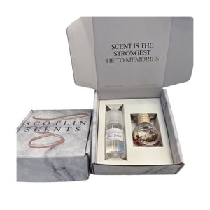 Op de afbeelding: Een witte doos met de tekst "SCOTLIN SCENTS" op de voorkant. De doos is open en bevat een klein flesje "Lilac Flowers" geurolie en een autoluchtverfrisser met een houten hanger. De doos staat op een wit oppervlak.