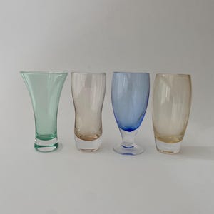Puede incluir: Cuatro vasos de chupito de vidrio en diferentes colores: verde, rosa, azul y amarillo. Los vasos son todos transparentes y tienen una superficie lisa.