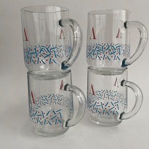 May include: Four clear glass mugs with handles are stacked. Each mug has a red letter "A" and a blue, red, and white confetti-like pattern. The mugs are transparent, allowing visibility of the contents.
