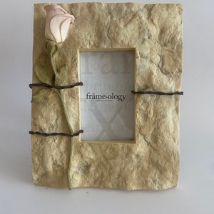 Könnte beinhalten: Ein beigefarbener Steinbilderrahmen mit einer geformten Rose und Stacheldraht-Akzenten. Der Rahmen hat einen weißen Passepartout und der Text "frame-ology" ist auf den Passepartout gedruckt.