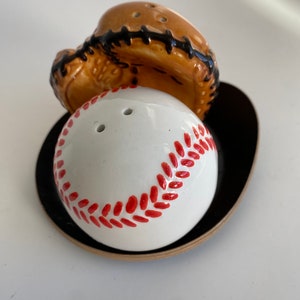 Puede incluir: Una pelota de béisbol de cerámica blanca con costuras rojas se asienta sobre una base negra y marrón. Un guante de béisbol de cerámica marrón se asienta encima de la pelota de béisbol.