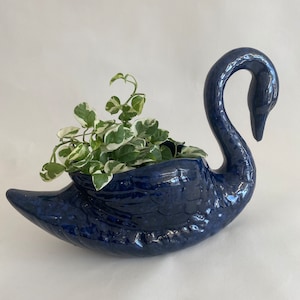 May include: A blue ceramic swan planter with a leafy green plant inside. The swan has a textured surface and a long neck.
