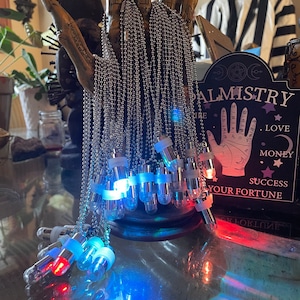 May include: A silver chain necklace with a variety of small, clear glass vials with colored lights inside. The vials are hanging from the chain and are arranged in a circle. The chain is hanging from a wooden hand with a palmistry chart in the background.