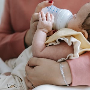 May include: A baby being fed from a white baby bottle by a person wearing a pink sweater. The baby is wearing a white and yellow outfit with a floral pattern.
