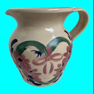 May include: A cream-colored ceramic pitcher with a heart-shaped design painted in pink, green, and blue. The heart is surrounded by pink flowers with blue centers.