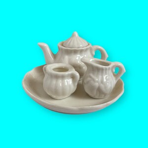May include: A white porcelain tea set with a teapot, creamer, and sugar bowl. The set is on a white saucer.