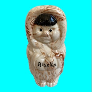 May include: A ceramic figurine of a person wearing a brown and white hooded parka. The figurine is painted with brown and white swirls and has the word "Alaska" written on the front.