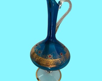 May include: A decorative blue glass pitcher with a clear handle and a gold rim. The pitcher features gold filigree and red accents. The base is light blue with a gold rim. The pitcher is approximately 12 inches tall.