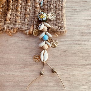 May include: A decorative keychain with a bohemian design. The keychain is made with seashells, gold-coloured coins, and a painted bead with a floral design. It is attached to a silver-coloured clasp and hangs against a natural fibre background.