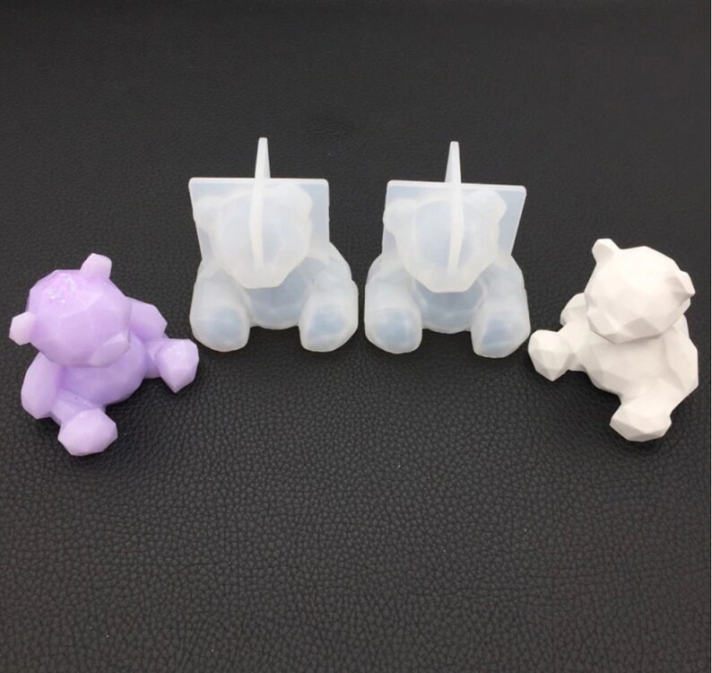 May include: A collection of bear-shaped items. There is a purple bear, two clear silicone molds, and a white bear. All bears have a geometric, faceted design. The items are arranged on a dark, textured surface.