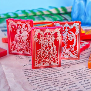 May include: Three red tarot card-shaped objects with gold-colored illustrations and text. The cards feature images of figures and the words "TEMPERANCE", "THE EMPRESS", and "THE WORLD". The cards are on a printed paper background.