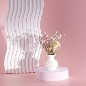 May include: A white, textured vase filled with dried flowers, set on a pink pedestal. A white, wavy, architectural element stands behind the vase against a pink backdrop. The flowers are a mix of white, pink, and beige.