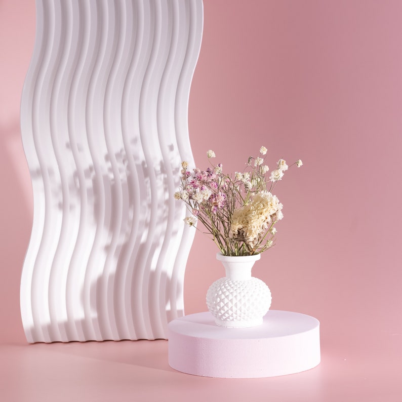May include: A white, textured vase filled with dried flowers, set on a pink pedestal. A white, wavy, architectural element stands behind the vase against a pink backdrop. The flowers are a mix of white, pink, and beige.