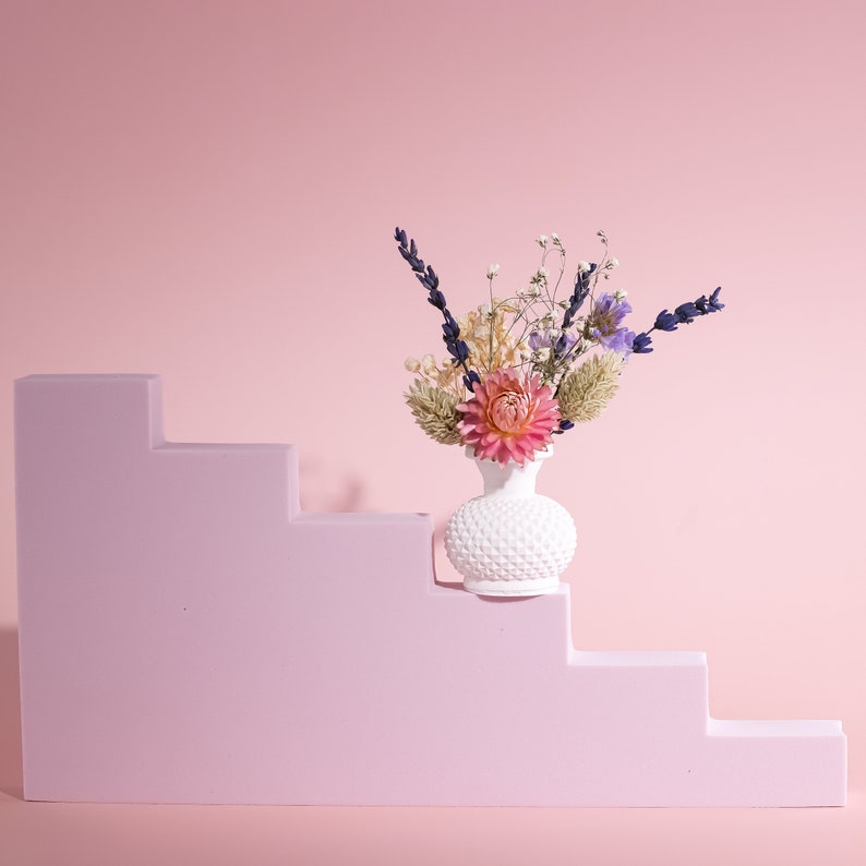 May include: A white, textured vase filled with dried flowers, including lavender and a pink dahlia, sits on a light purple staircase. The background is a solid pink, creating a soft, minimalist aesthetic. The vase has a unique, dotted pattern.