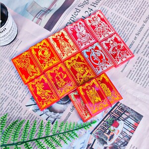 May include: A collection of red tarot cards with gold and white illustrations. Each card features a unique design, including figures and symbols. The cards are arranged on a newspaper background, creating a visually appealing display.