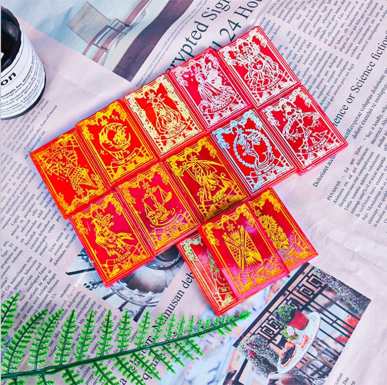 May include: A collection of red tarot cards with gold and white illustrations. Each card features a unique design, including figures and symbols. The cards are arranged on a newspaper background, creating a visually appealing display.