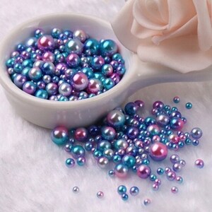 Puede incluir: Una colecci&oacute;n de perlas iridiscentes en tonos azules, morados y rosas. Las perlas est&aacute;n dispersas sobre una superficie blanca y en un peque&ntilde;o cuenco blanco. Una suave rosa rosa est&aacute; en el fondo.