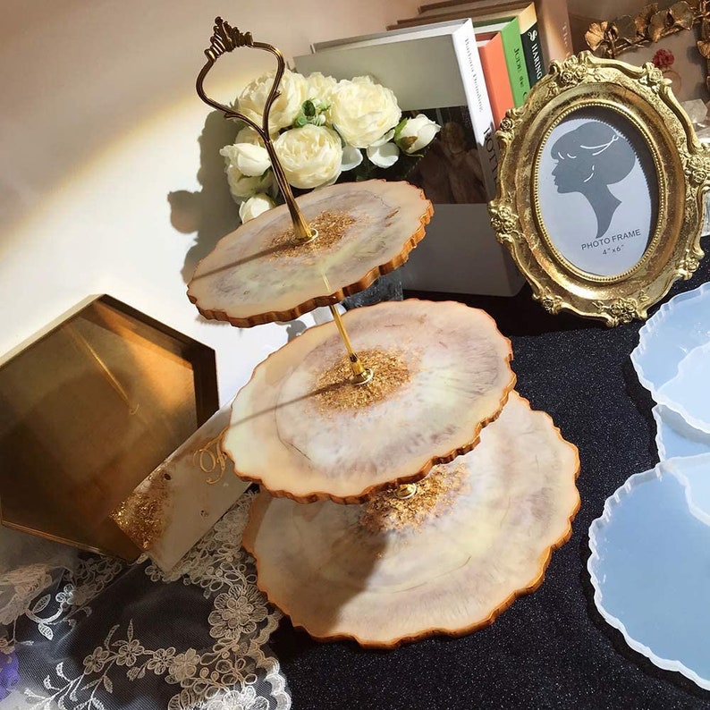 May include: A three-tiered serving stand with a gold handle and support. The tiers are round with a light beige and gold resin design. The stand is displayed with a gold framed photo and white flowers.