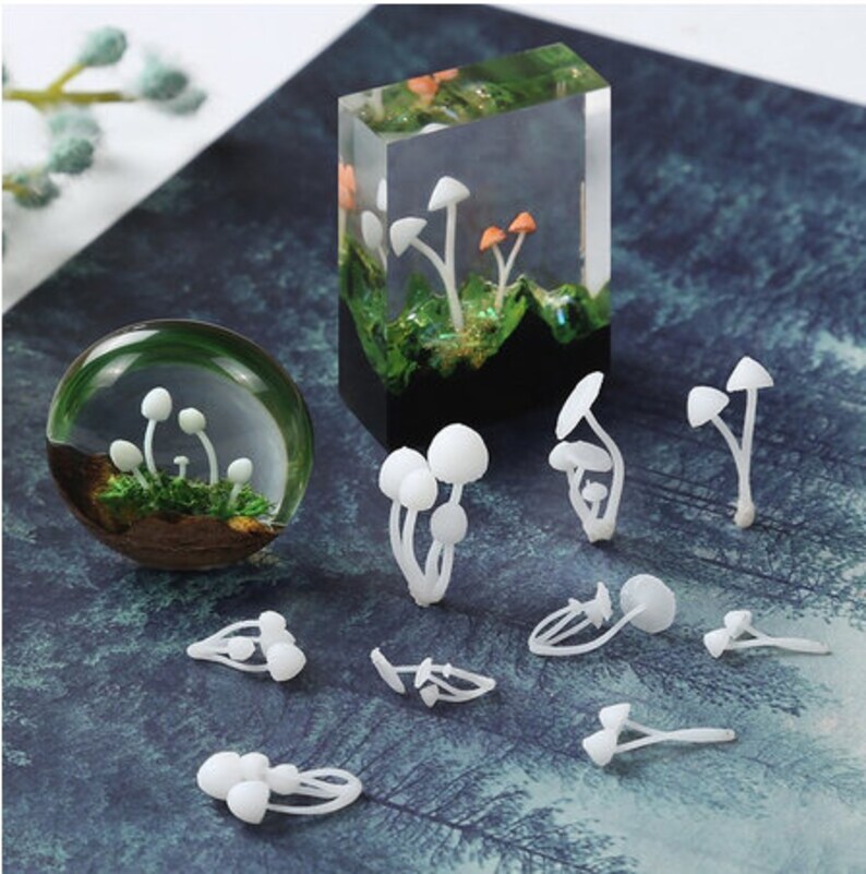 May include: A selection of mushroom-themed art pieces. Features a clear resin block and a glass sphere, both with miniature mushrooms and greenery. Several white mushroom figurines are also displayed, showcasing a whimsical, nature-inspired theme.