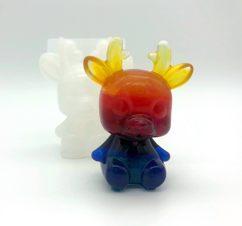 May include: A colorful resin deer figurine with a gradient of red, orange, and blue hues. The deer has yellow antlers and a cute, rounded design. A white deer-shaped mold is in the background.