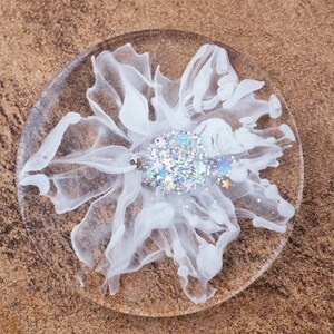 May include: A clear, round coaster with a white, abstract floral design. The center of the flower features iridescent glitter and small star-shaped accents. The coaster is made of resin.