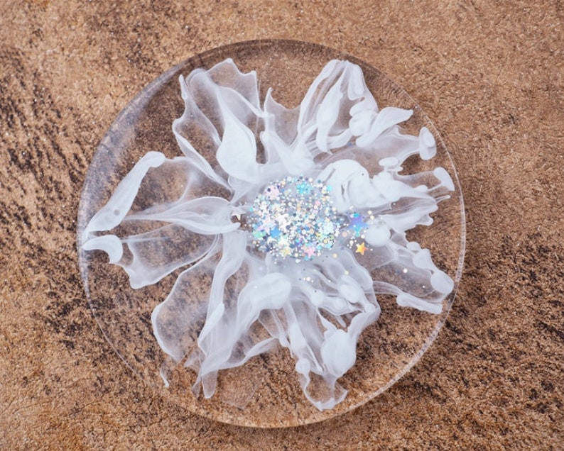 May include: A clear, round coaster with a white, abstract floral design. The center of the flower features iridescent glitter and small star-shaped accents. The coaster is made of resin.