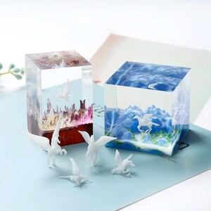 May include: Two clear resin cubes with miniature scenes inside. One cube features a cityscape with a white pegasus, the other a mountain landscape with a pegasus. Several white pegasus figurines are in front.